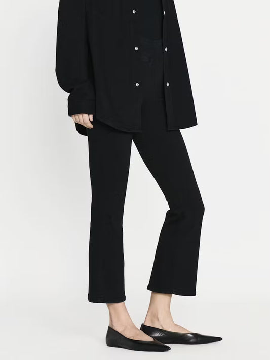 A person in a black long-sleeve shirt and Frame's Jetset Crop Mini Pintuck Pants stands against a plain background. They sport black flats, their right hand resting at their side. Their face is not visible in the image, highlighting the elegance of the high-rise silhouette.