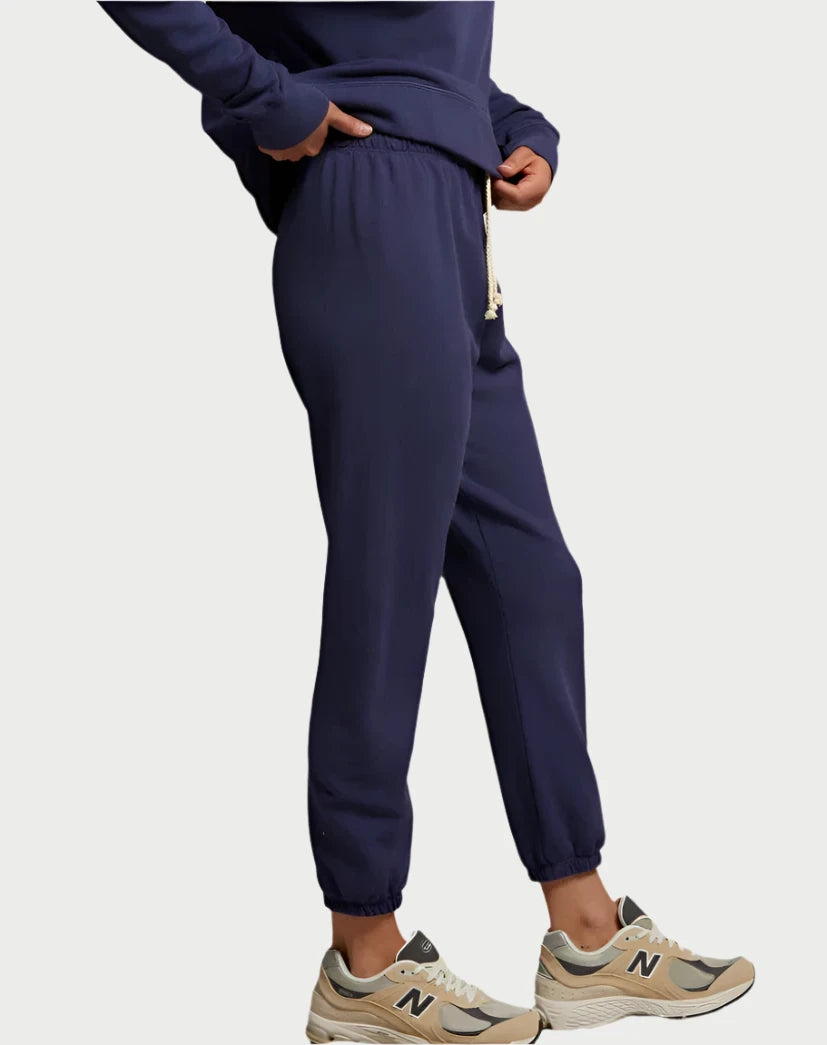 A person is modeling the Johnny Sweatpant in Dusk Blue from perfectwhitetee, featuring an elastic waistband and drawstring, crafted from French terry cotton. They pair these pants with beige and green sneakers, while a navy sweatshirt is visible over a crisp white tee against a minimalist California-style plain white backdrop.