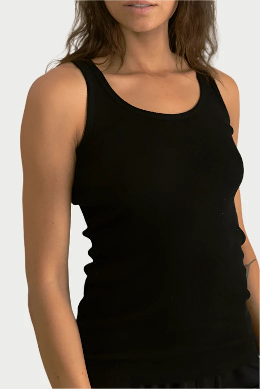 A person with long, wavy brown hair and freckles stands in front of a neutral backdrop. They are wearing the perfectwhitetee Jagger Layering Tank, a black, sleeveless top made from soft cotton, looking to the side with a calm expression. This versatile wardrobe piece is perfect for any occasion.