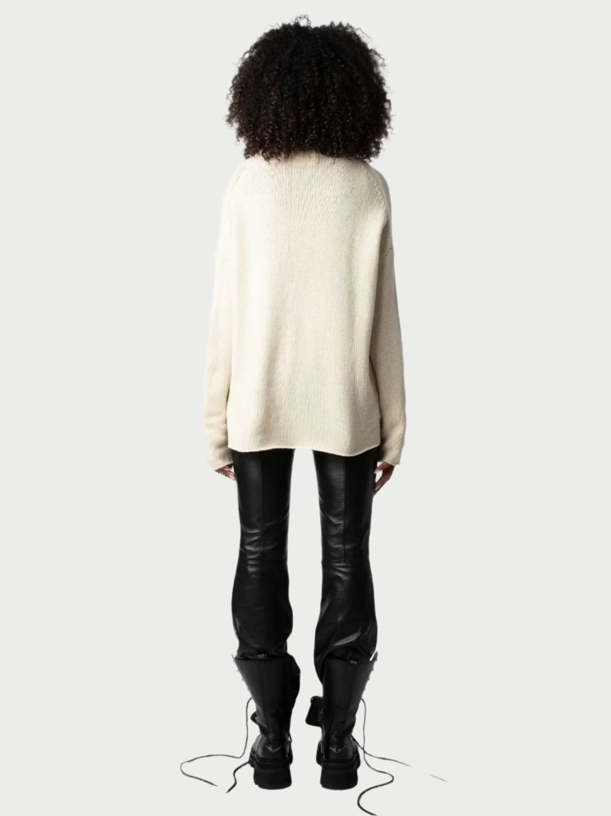 A person with curly hair is standing facing away, wearing a long, loose-fitting Peace Sweater by Zadig & Voltaire in light yellow, black leather pants, and black combat boots. The laces of the boots are undone and trailing on the floor. The background is a plain light gray.