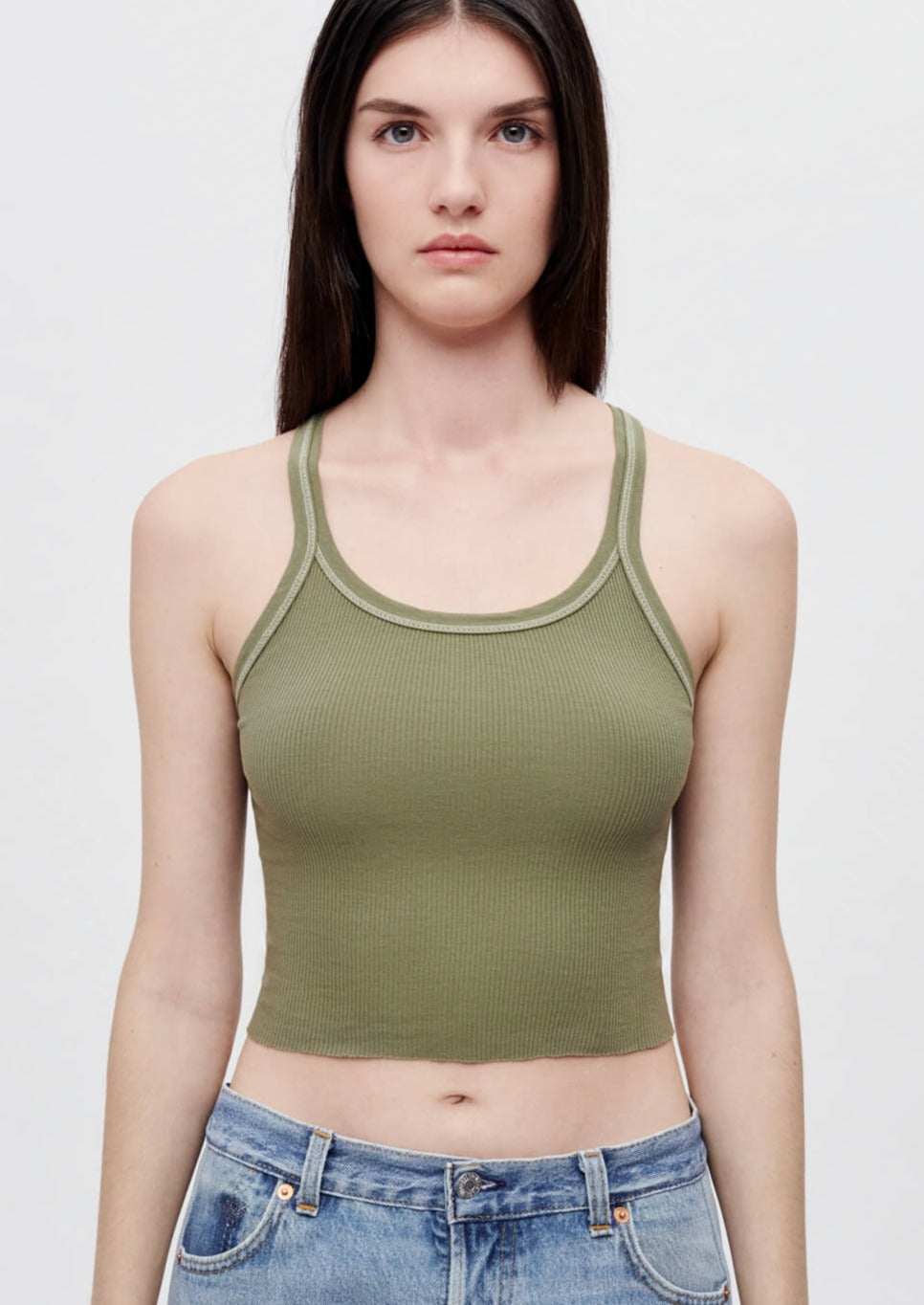 A young woman with long dark hair is wearing a sleeveless RE/DONE Cropped Rib Bayleaf Tank in olive green and light blue jeans. The cropped length top accentuates her outfit as she stands against a plain white background, looking directly at the camera with a neutral expression.