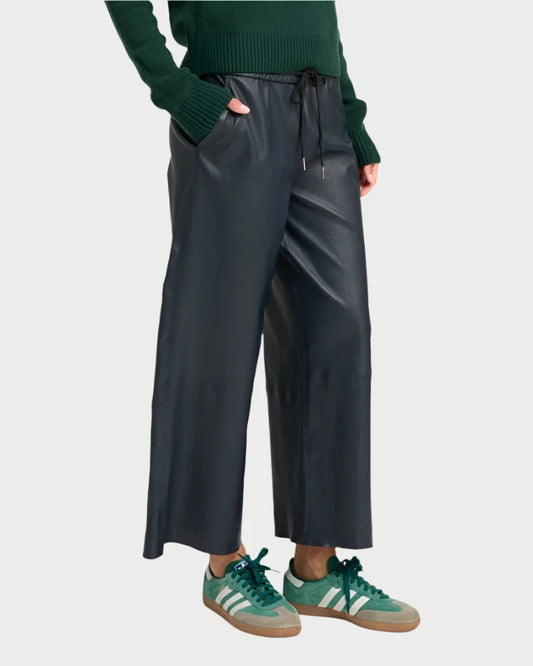 A person wearing a dark green sweater and SPRWMN's Cropped Baggy Leather Pants in Midnight, showcasing a stylish, cropped silhouette. They are also wearing green and beige sneakers against a plain light grey background.