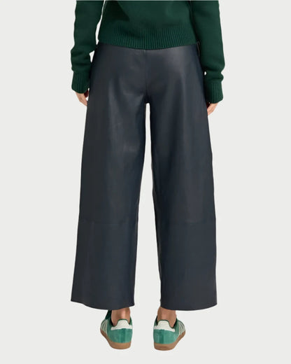 A person is wearing SPRWMN's Cropped Baggy Leather Pants in Midnight paired with green sneakers and a green sweater. The image, taken from behind, emphasizes the cut and fit of the leather trousers.