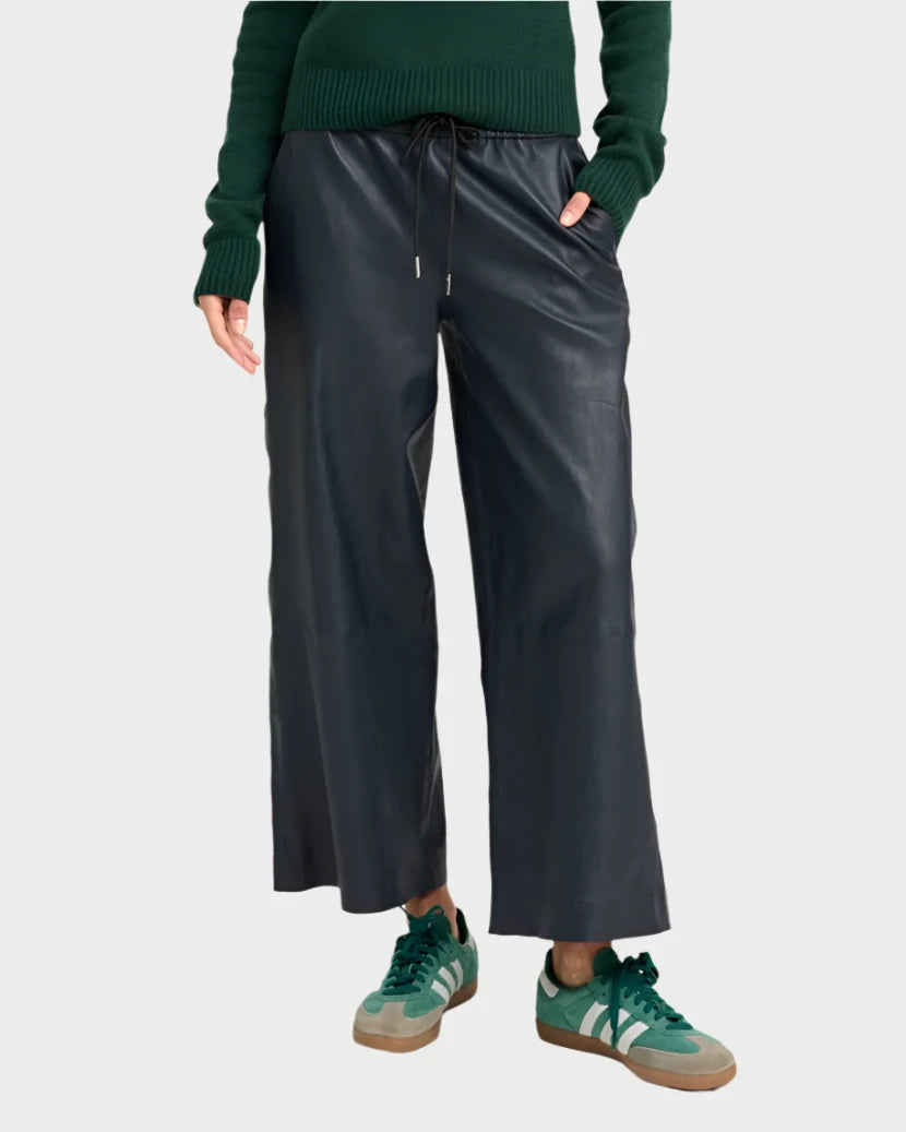 A person wearing a dark green sweater, SPRWMN's Cropped Baggy Leather Pants in Midnight, and green sneakers with white accents exudes a casual yet stylish vibe. The individual stands with one hand in their pocket against a light background, showcasing a chic, cropped silhouette.