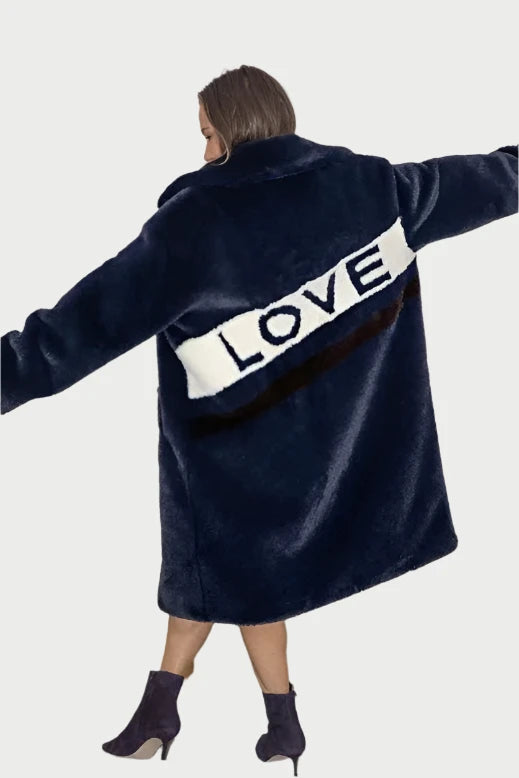 A person wearing a dark blue Mr Greys Love Coat by ANGEL WINGS, featuring the word "LOVE" written on the back in white and black, stands with arms outstretched like angel wings. They are indoors, and behind them are a light-colored curtain and a low table with yellow objects.