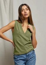 Load image into Gallery viewer, A woman with long brown hair is standing in front of a plain background. She is wearing the Margot- Safari sleeveless, V-neck, green top by perfectwhitetee and blue jeans—perfect for everyday wear. Her left hand is placed on her waist, and she has a relaxed expression on her face.