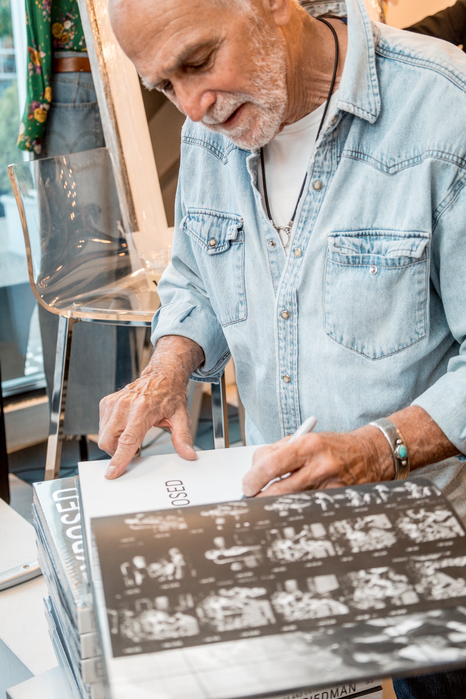 An elderly man with a white beard and wearing a light denim shirt autographs a copy of "EXPOSED - The Lost Negatives" by Michael Friedman Photography. He stands beside a clear acrylic chair, while a colorful piece of fabric, reminiscent of Rock n' Roll history from the 1960's music industry, is partially visible in the background.