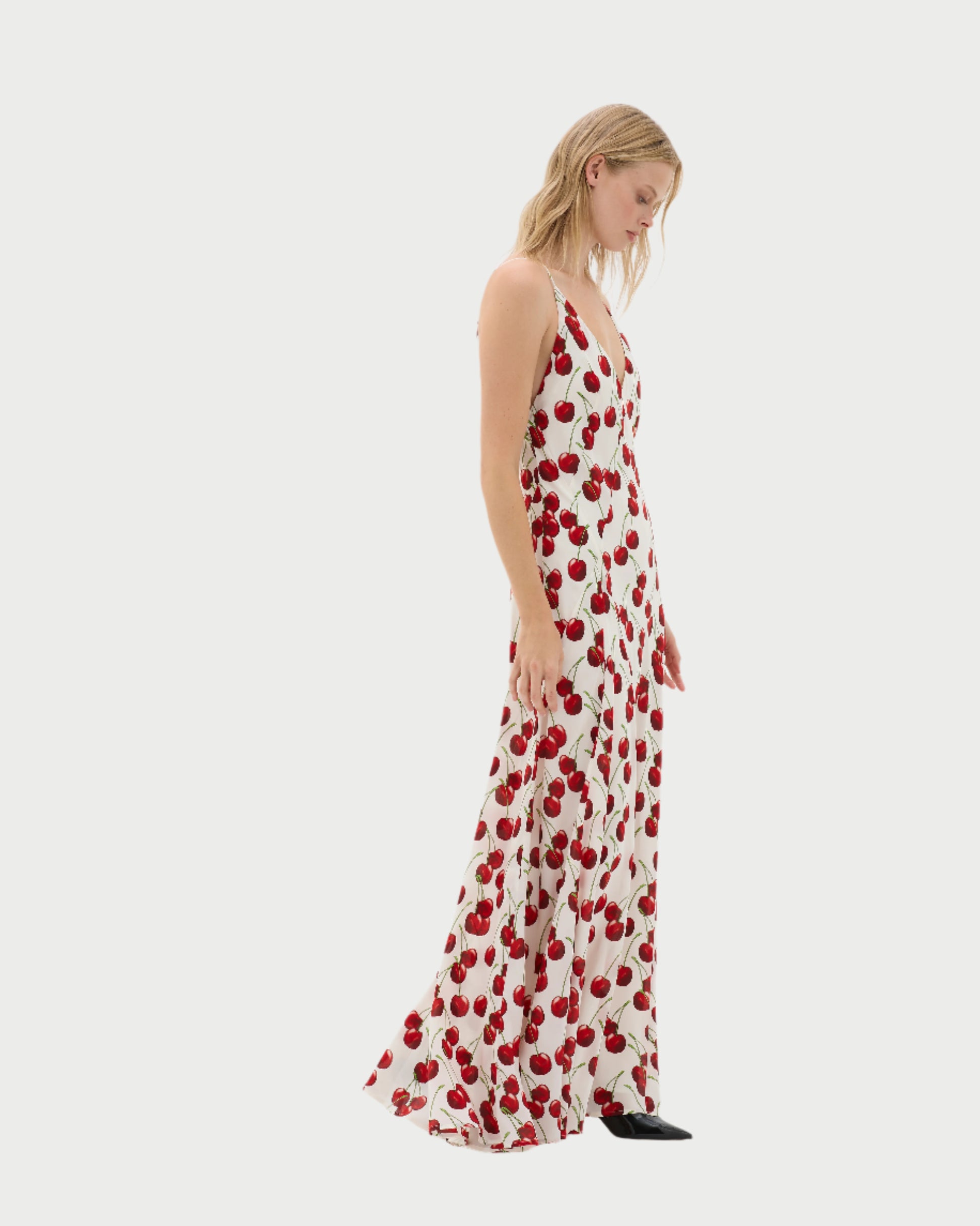 A person in a MUMA Altair Cherries Dress, featuring a cherry print, V-neckline, and thin straps, stands sideways against a plain background with their head slightly tilted down as the maxi dress flows gracefully to the floor.