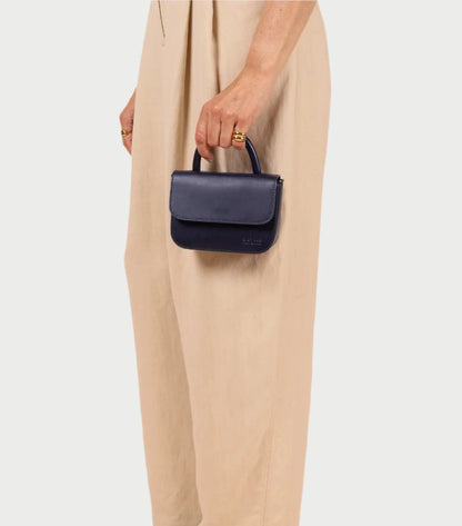 Someone in beige pants holds a stylish O MY BAG Nano Bag in dark blue with a short handle, perfectly complementing the minimalist white backdrop.