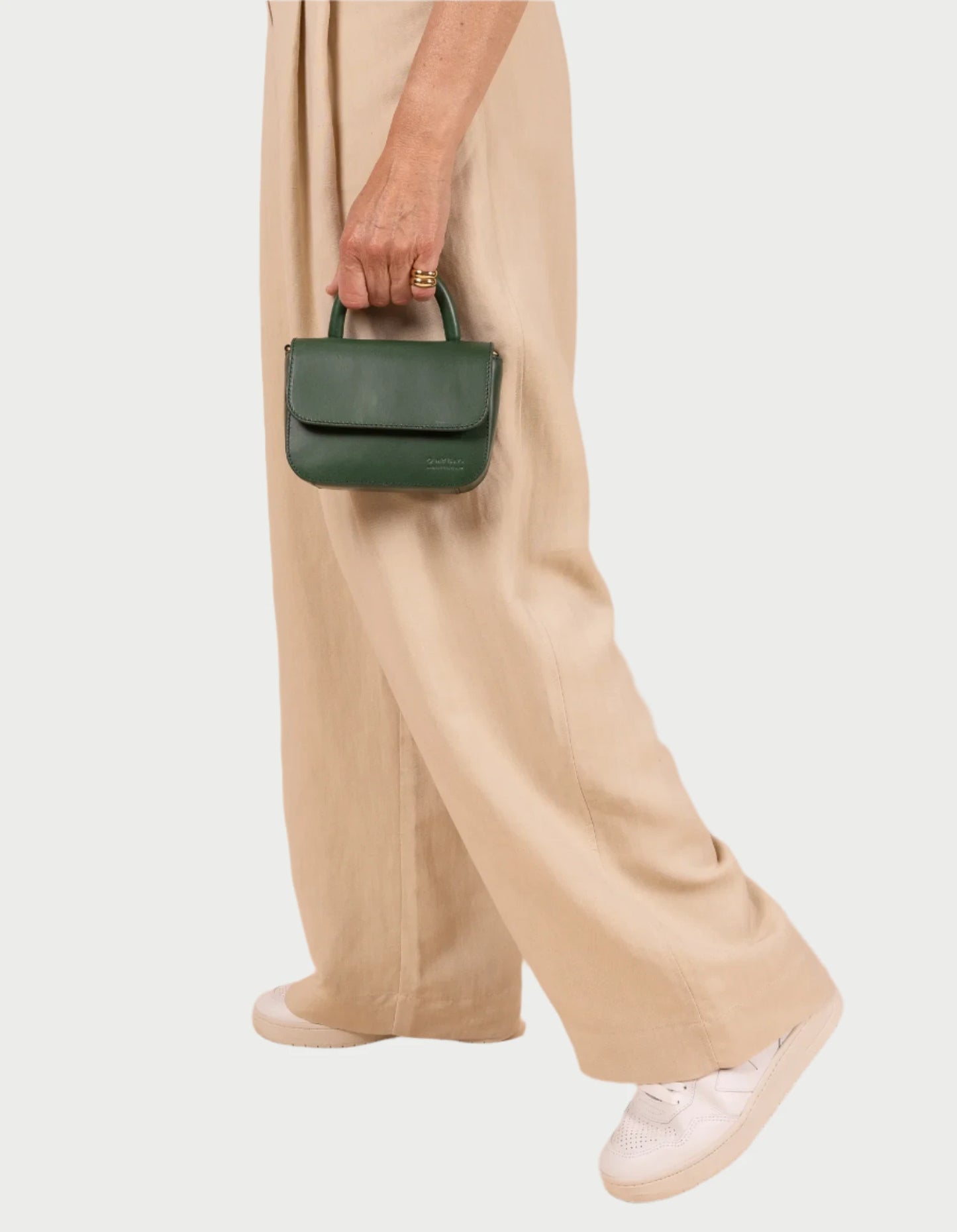 A person with beige pants and white sneakers carries an O MY BAG Nano Bag, small and green with a crossbody strap. Their minimalist outfit is chic, accented by the stylish bag against a plain white background.