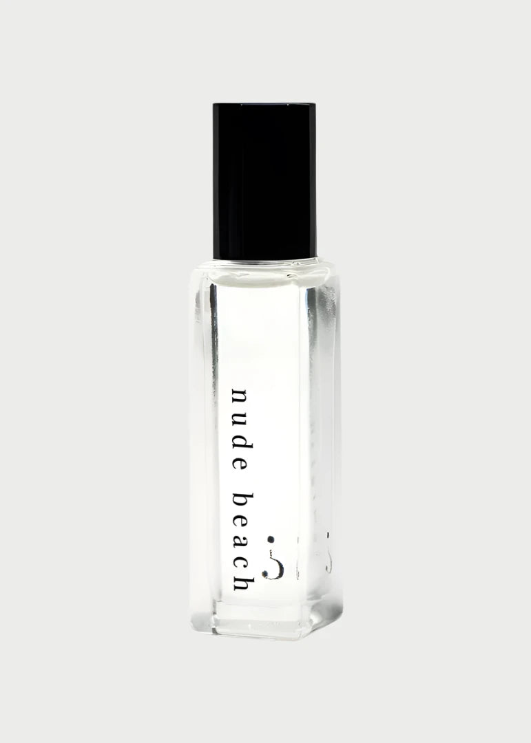 A Riddle Oil - Nude Beach 8ml bottle, made of clear glass and sealed with a black cap, contains a transparent liquid. The label features the words "nude beach" in lowercase letters. The design is minimalist and sleek, showcasing the trending Nude Beach scent by Riddle Oil.