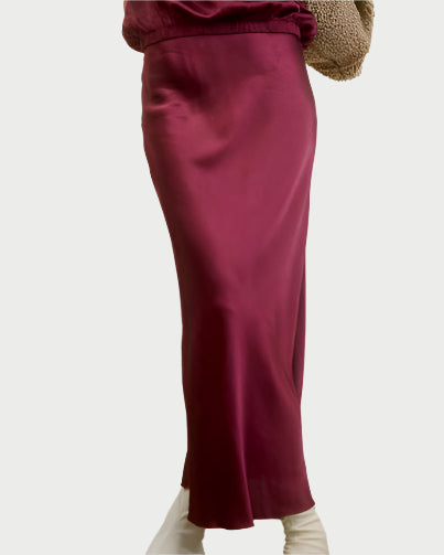 A person stands against a plain background, wearing an AqC O'Darling Skirt in wine-colored satin paired with a beige textured sweater. Their lower legs are visible in white shoes, accentuating the ankle-grazing elegance of their chic ensemble.