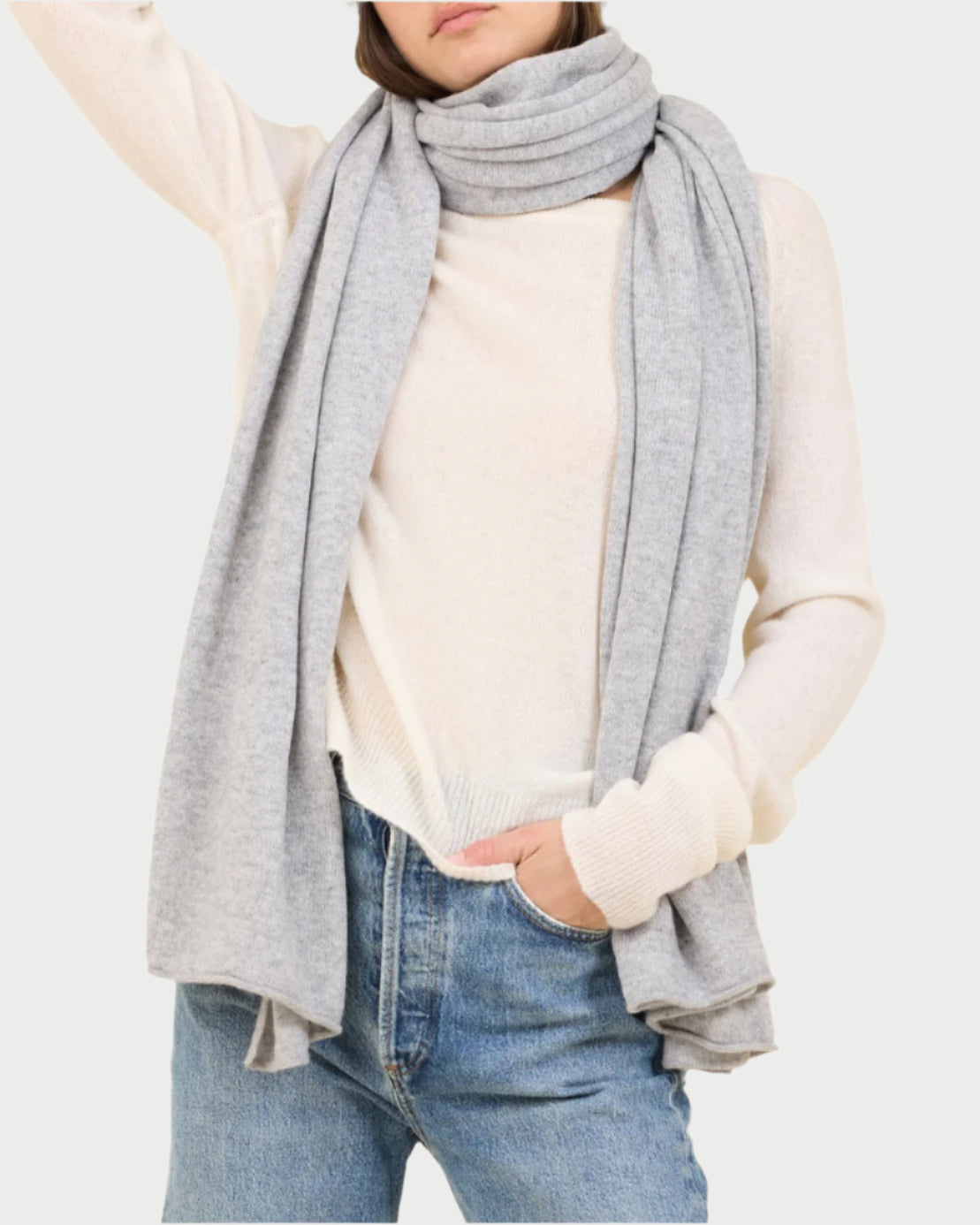 Against a beige background, a person effortlessly exudes luxury in blue jeans and a white sweater, accessorized with One Grey Day's light gray Cashmere Travel Scarf and matching beanie. They stand confidently with a hand on their hip.