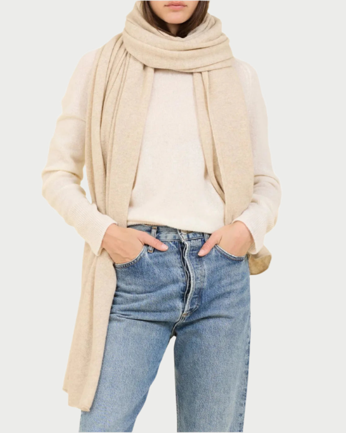 A person stands against a light beige background wearing a luxurious beige knit hat, One Grey Day Cashmere Travel Scarf, cream sweater, and blue jeans. Their hands are tucked into their jeans pockets.