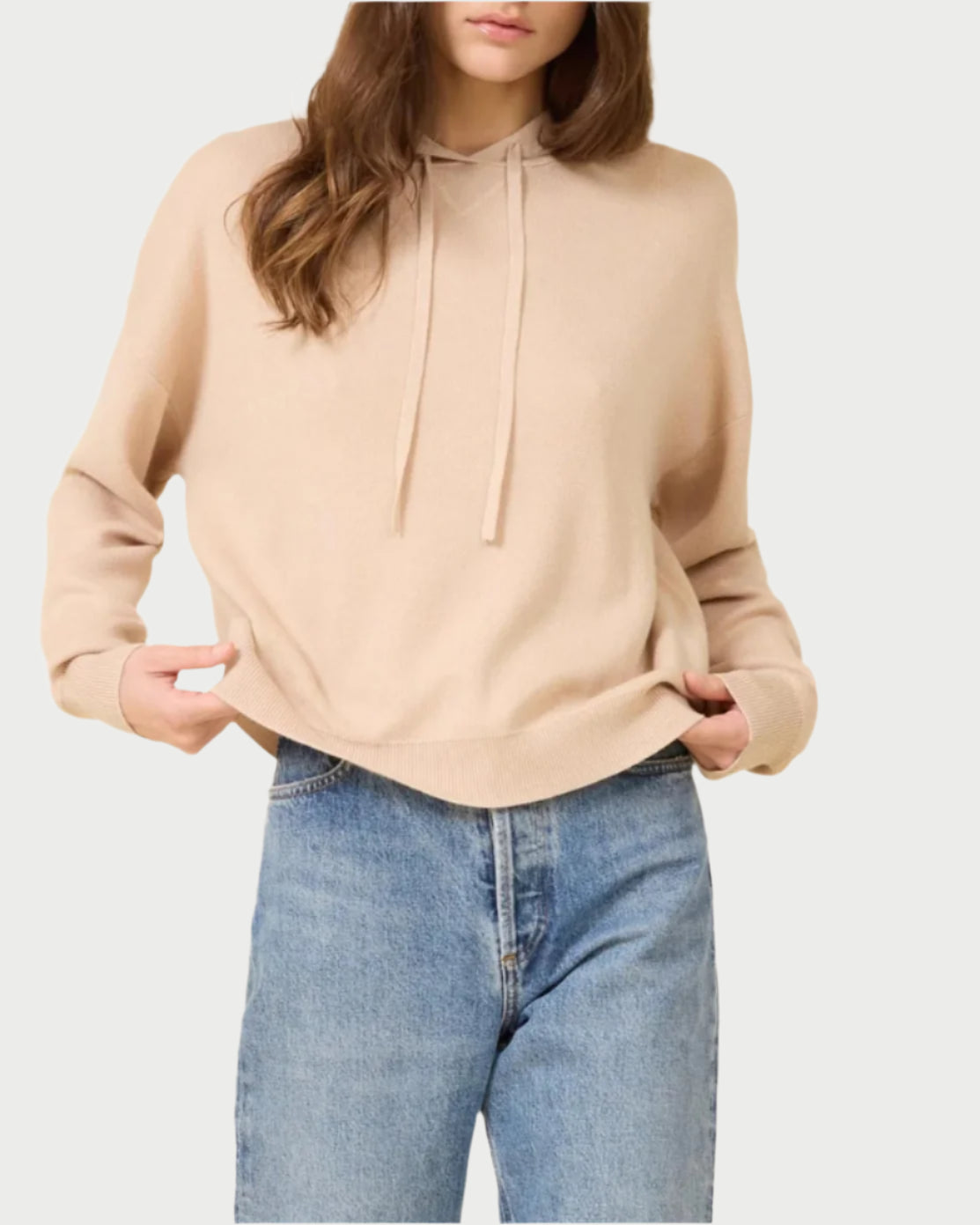 A person adjusts the hem of a super soft Warwick Hoodie in Latte by One Grey Day, paired with light blue jeans, against a plain white background. The head is partially out of frame.