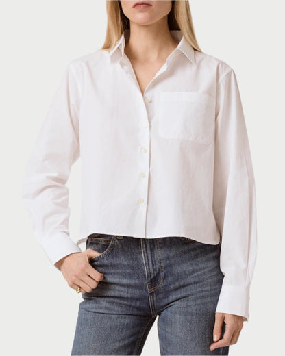 A person with long, light-colored hair wears a chic Coco Cropped Shirt by T. Line—a slightly cropped, 100% cotton white shirt with a chest pocket—and blue jeans, standing with one hand in their pocket.