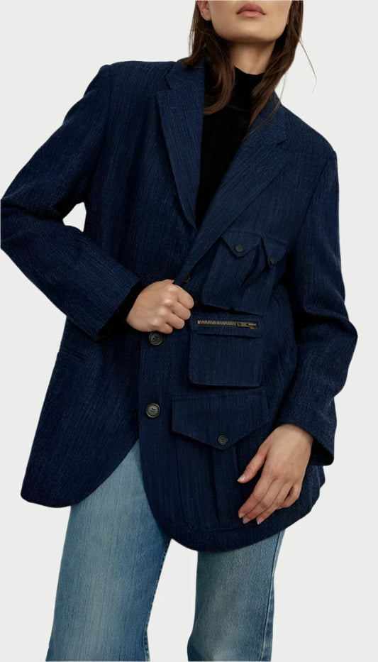 A person is styled in the Surplus Blazer from Smythe, showcasing its oversized silhouette in Indigo Blue, which features a front zipper pocket and large buttons. It is elegantly layered over a black top and paired with light blue jeans. The head is partially cropped out of the image.