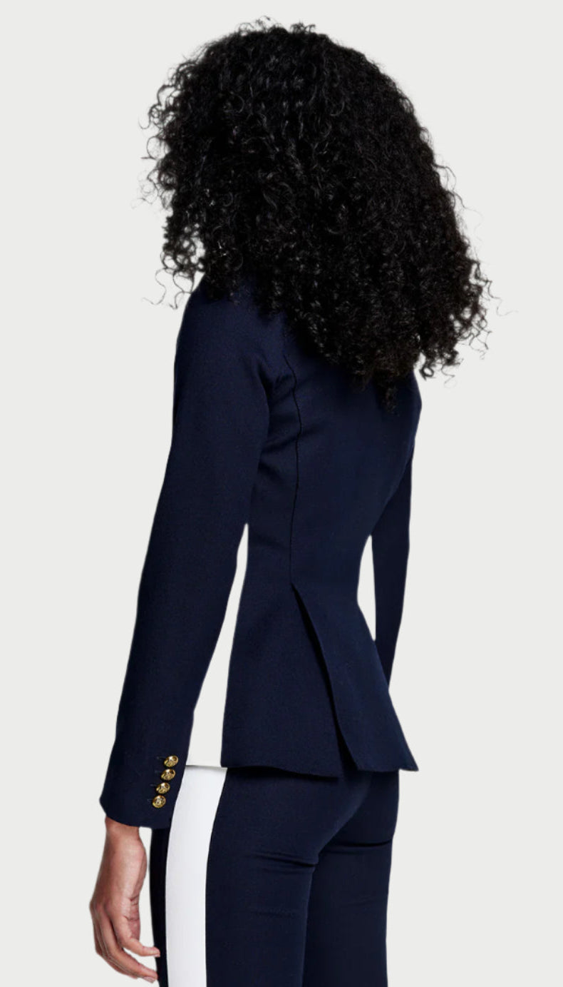 A person with curly hair wears the Smythe Patch Pocket Stripe Blazer in Deep Navy, featuring gold buttons and matching pants with a contrast tux stripe, turned away from the camera against a light gray background.