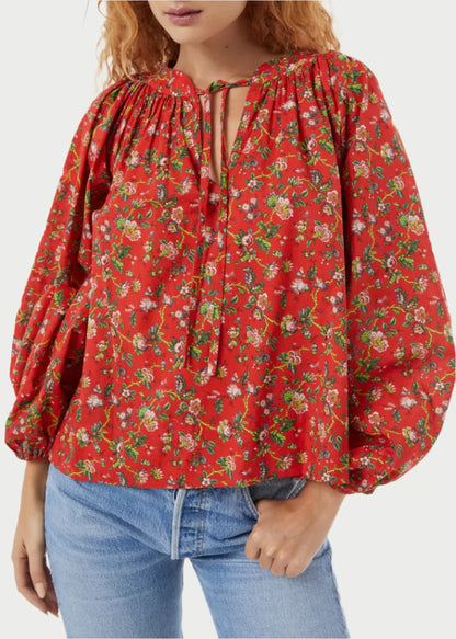 A woman with curly red hair is wearing Rhode's Red Flowered Poet Top, featuring puffy sleeves and a tie at the neckline, crafted from organic cotton. She stands against a plain white background with one hand in the pocket of her light blue jeans, emphasizing a timeless silhouette.