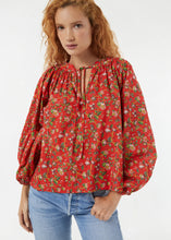 Load image into Gallery viewer, A woman with curly red hair is wearing Rhode&#39;s Red Flowered Poet Top, featuring puffy sleeves and a tie at the neckline, crafted from organic cotton. She stands against a plain white background with one hand in the pocket of her light blue jeans, emphasizing a timeless silhouette.