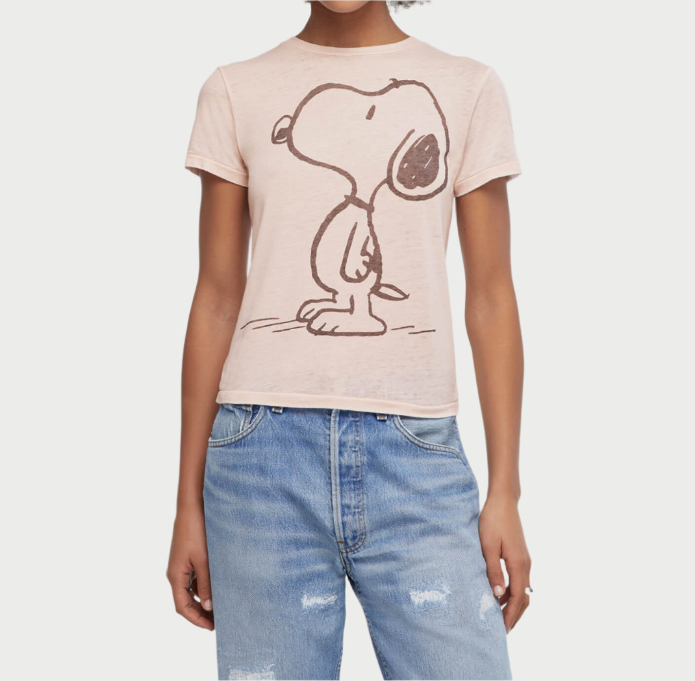 A person wearing a RE/DONE Dahlia Snoopy Tee in light pink, featuring a sketch of standing Snoopy, paired with light blue ripped jeans.