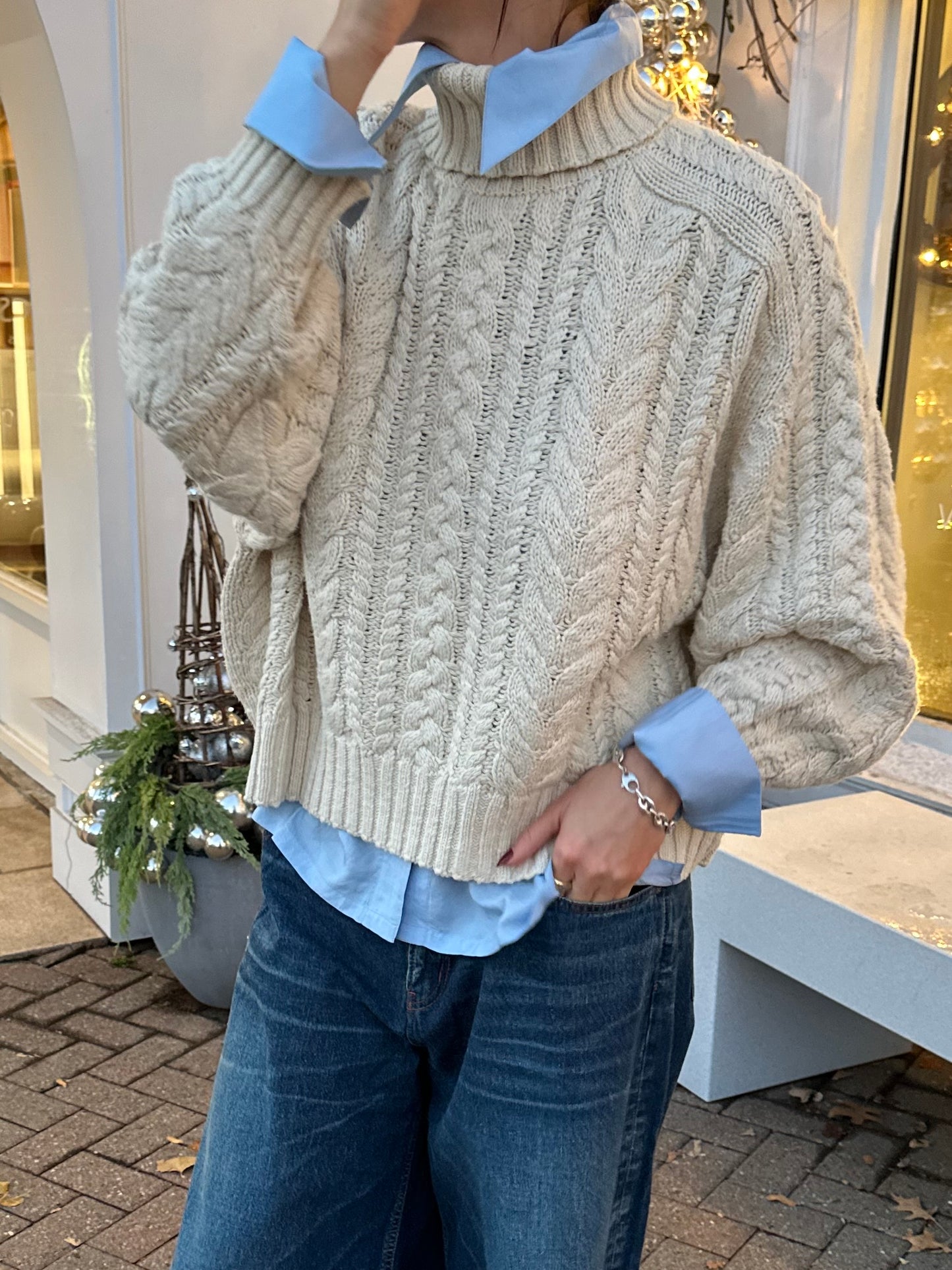 A person in a cozy cream cable-knit sweater, layered over an Alix of Bohemia's Dolly Bluejay Silk Shirt featuring oversized cuffs, paired with jeans. They are standing outdoors on a brick pathway, with a decorative plant and festive background visible.