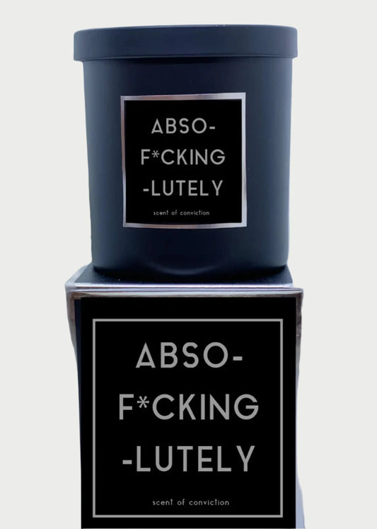 A black 9oz candle from Rude Dude with a label that reads "ABSO-F*CKING-LUTELY, scent of conviction" sits on a matching black box adorned with the same label. In the blurred background, a silver hand sculpture is visible, holding up its middle finger. Made in USA.