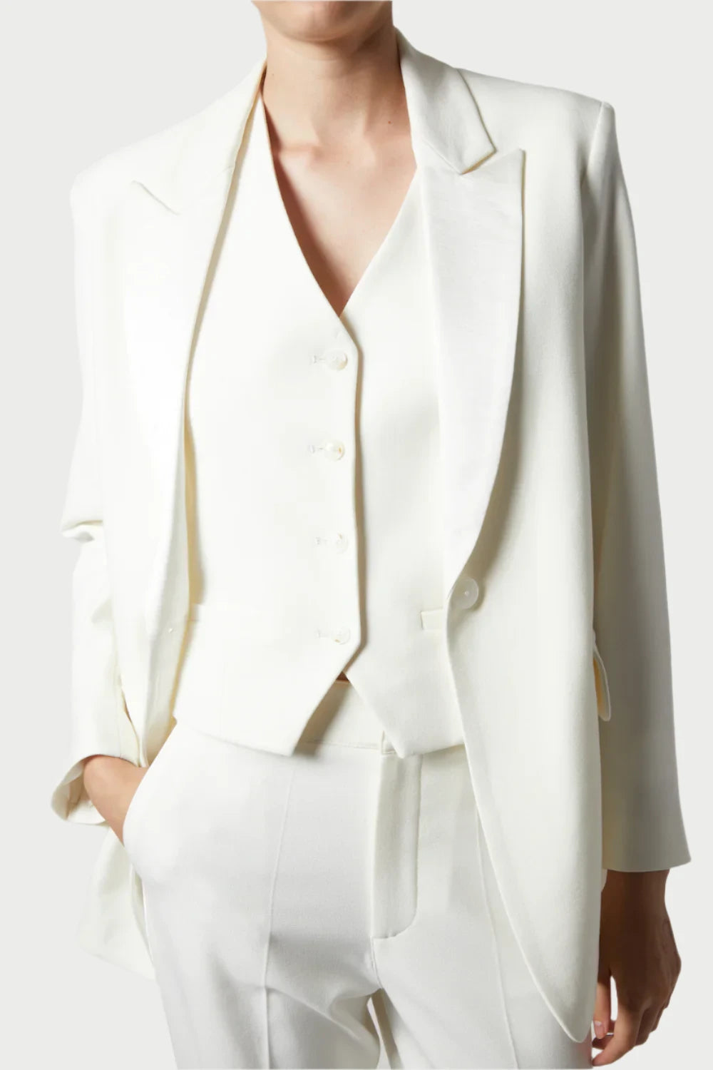 A person is dressed in a chic all-white ensemble featuring the Dylan Tuxedo Jacket in Ivory by Saint Art NY. The suit includes a tuxedo blazer, vest, and pants, showcasing a sleek and tailored appearance with charmeuse satin details. The individual poses gracefully with one hand in their pocket set against a neutral backdrop.