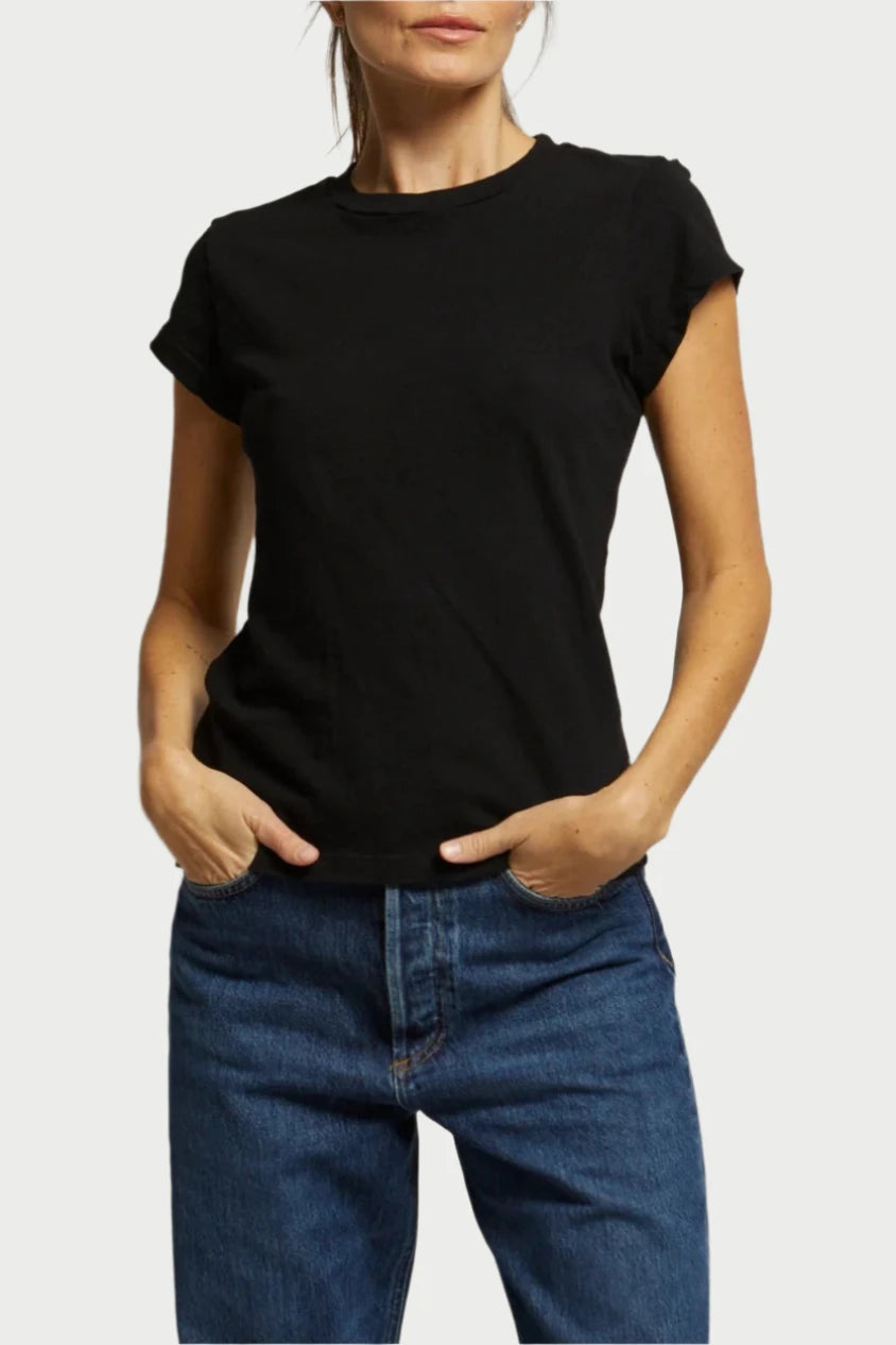 A person with dark hair in a ponytail stands against a plain background. They are wearing the Sheryl Tee by perfectwhitetee and blue jeans, their hands in their pockets. With a neutral expression, they gaze at the camera, casually showcasing the ideal tee for any occasion.