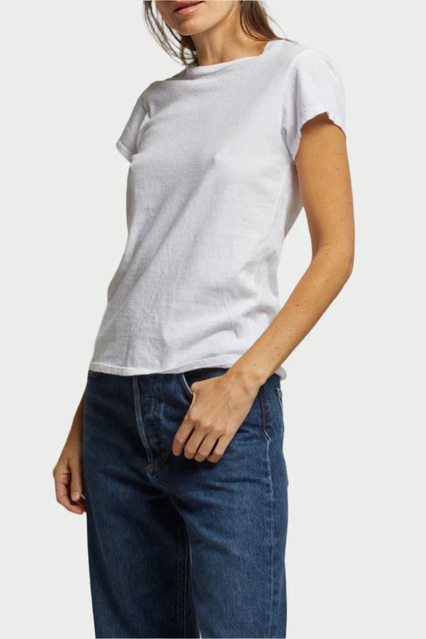 A person with long hair stands against a plain, light-colored backdrop. They are wearing the Sheryl Tee by perfectwhitetee and blue jeans, with their right hand resting lightly on their hip. Their expression is relaxed, and they are looking towards the camera.