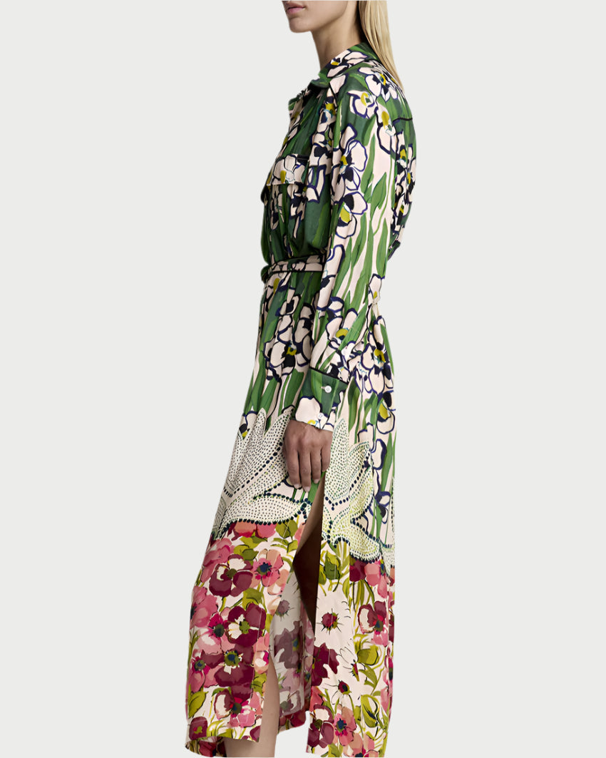 A person with long blonde hair is wearing Smythe's Tailored Shirt Dress, flaunting its garden-like Eco Vero-certified fabric in a mix of green, purple, white, and pink. They stand sideways in brown slip-on shoes, enhancing the dress's vibrant floral design.