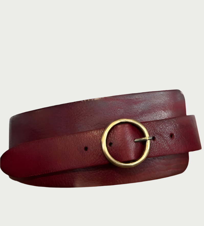 The Wide Contour Belt by Motif56, a dark red leather accessory with a simple gold round buckle, rests coiled on a plain background. Its smooth texture and high-quality craftsmanship evoke 70's boho-chic vibes.