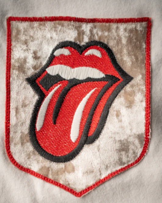 The Madeworn Rolling Stones Patch Pocket Tee features a vintage rock vibe with an embroidered velvet patch showcasing the iconic red lips and tongue design, set within a shield-shaped border for a timeless and eye-catching look.