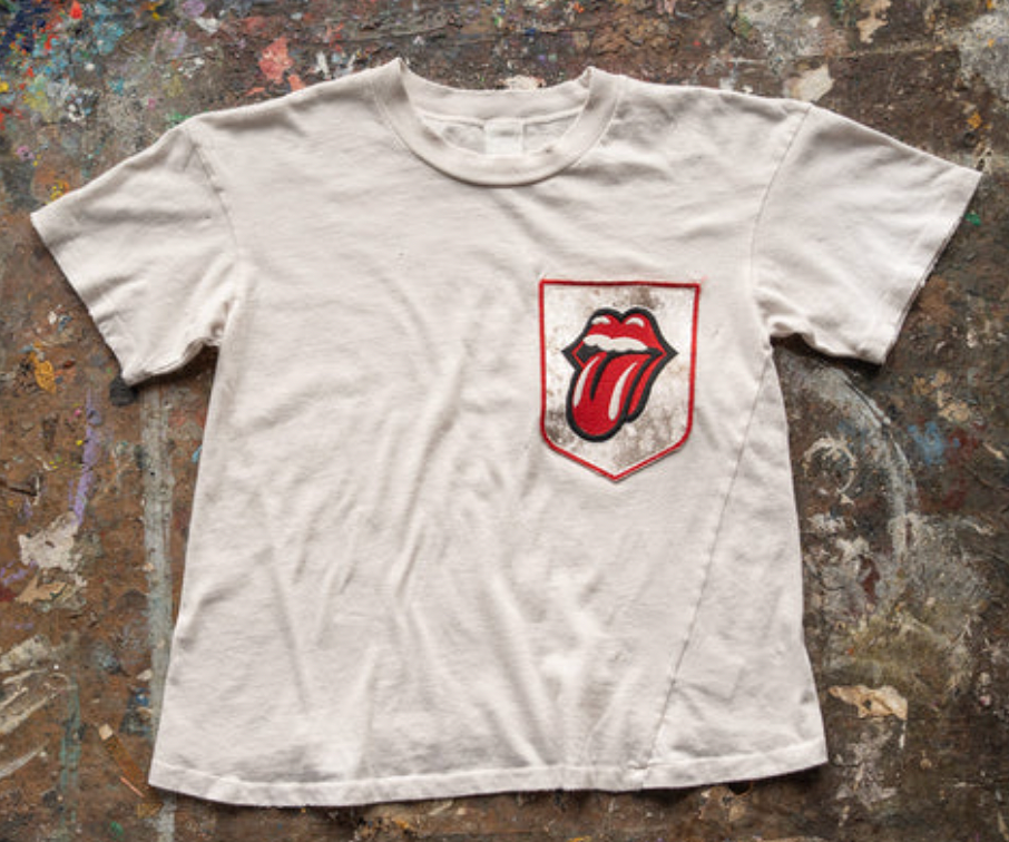 The Madeworn Rolling Stones Patch Pocket Tee is a vintage white rock T-shirt with the iconic red tongue and lips logo on the chest pocket, laid on a paint-splattered surface capturing classic rock style.