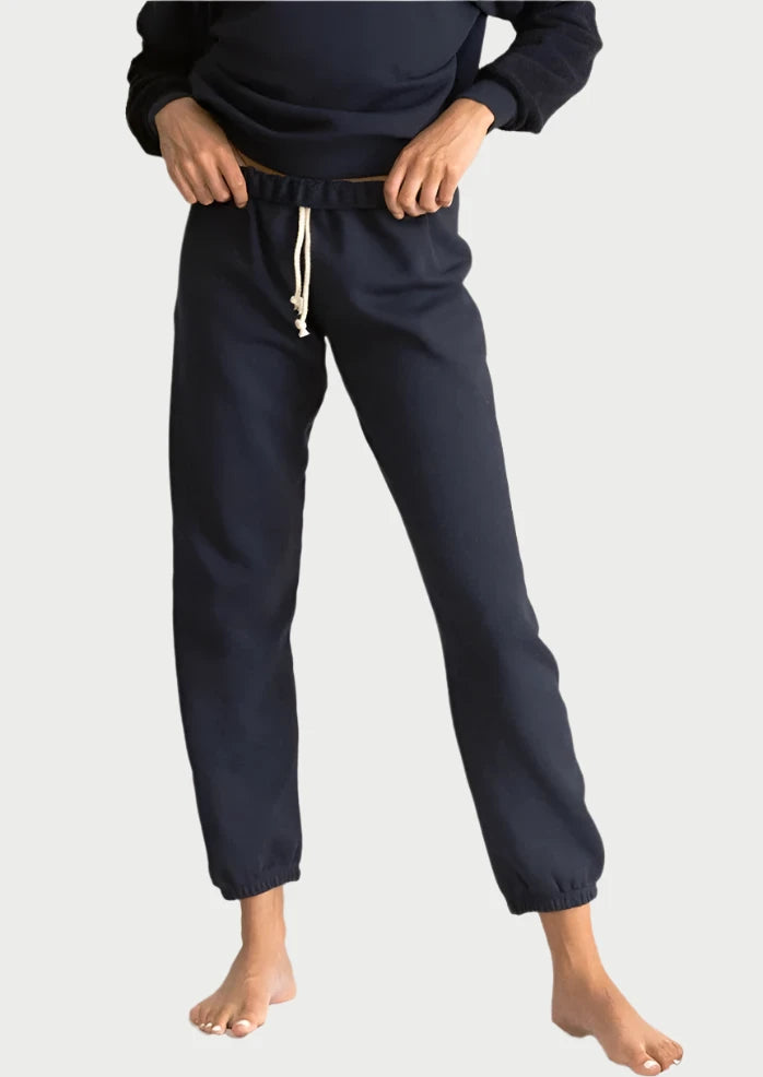 A person wearing a matching dark blue sweatshirt and high-waisted Stevie Sweatpant from perfectwhitetee stands barefoot on a fabric backdrop. Their hands are tucked into their joggers' pockets, and the hidden drawcord waistband is visible. The outfit looks comfortable and casual.