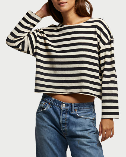 Standing against a light fabric background, a person wears the Taylor Striped Cotton Tee by perfectwhitetee, paired with loose blue jeans and red sneakers. They have long hair and rest one hand on their head, proudly showcasing a California-made piece.