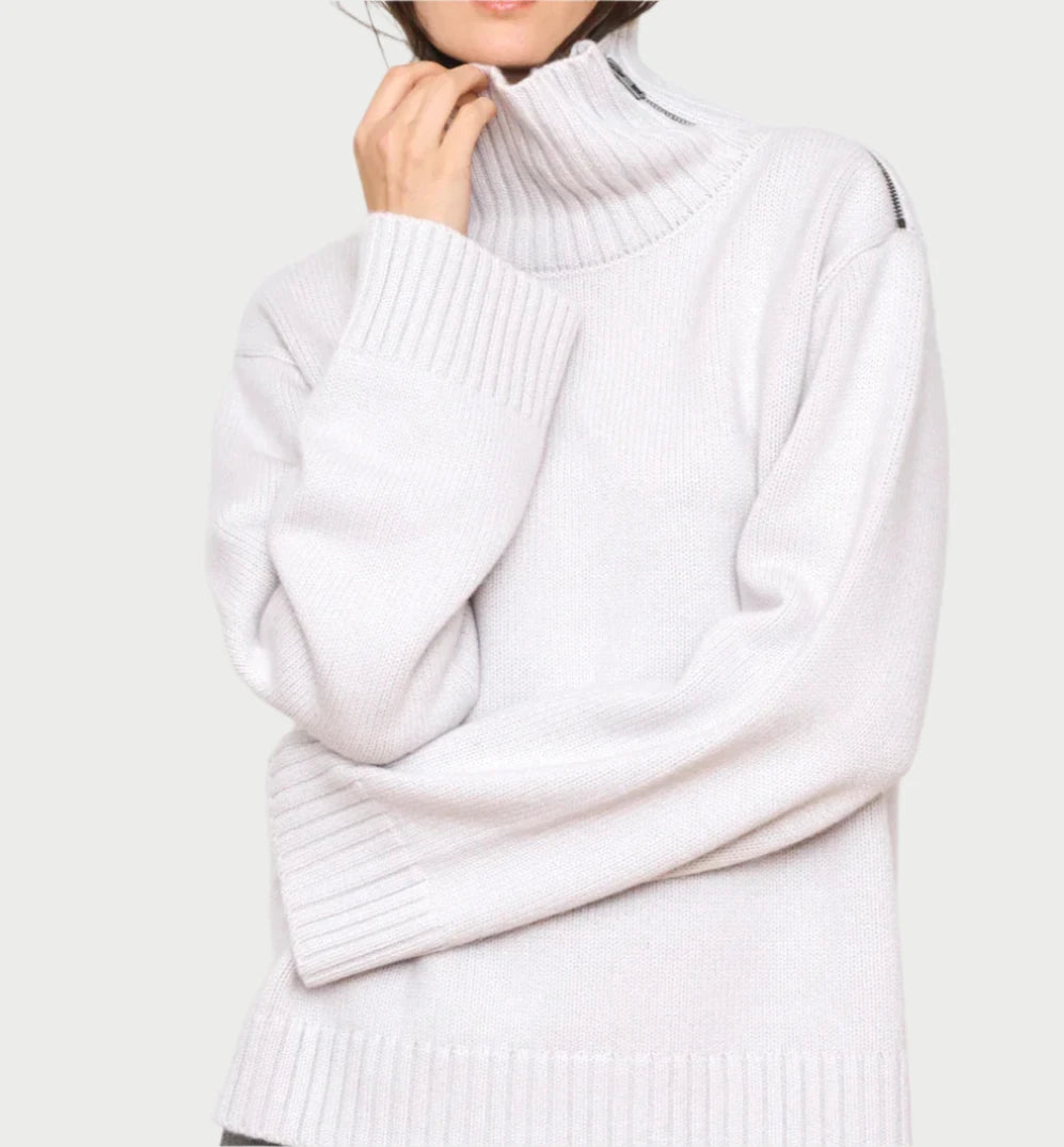 A person wearing a light gray, oversized Bowery Knit Sweater from WE-AR4, made with luxury deadstock materials, effortlessly adjusts the collar. The sweater boasts ribbed cuffs and hem against its simple light gray design. The person is only partially visible from the chest up.