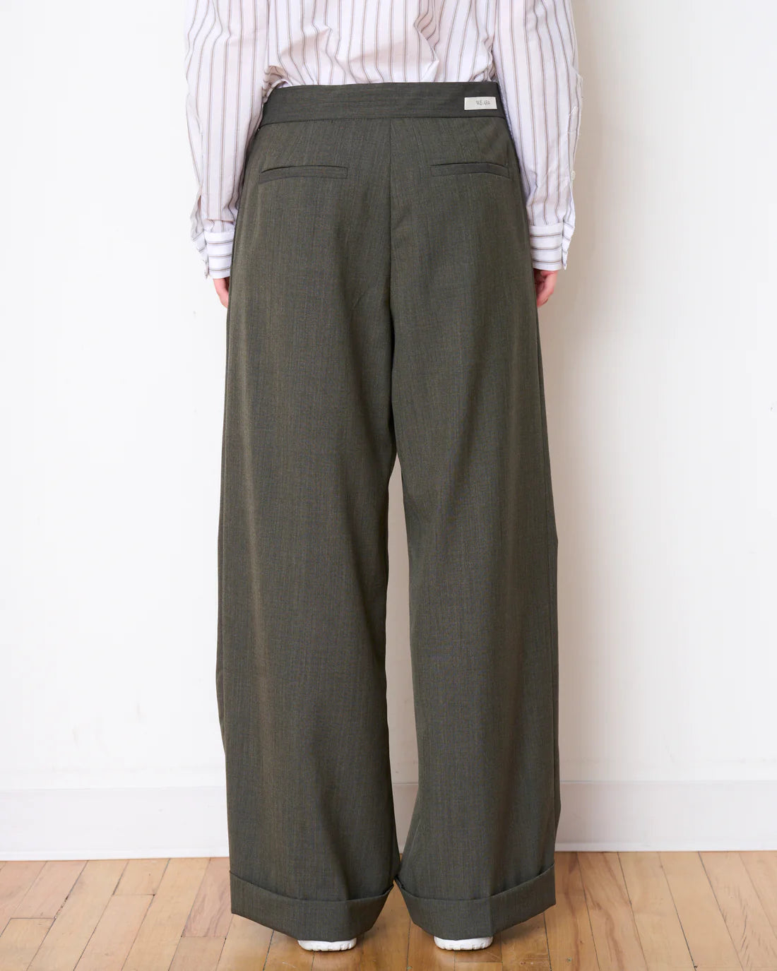 A person is positioned against a white wall, wearing a striped shirt and WE-AR4's Lounge Trousers made from upcycled materials. These wide-legged, olive green pants include two back pockets. The individual faces away from the camera while standing on a wooden floor.