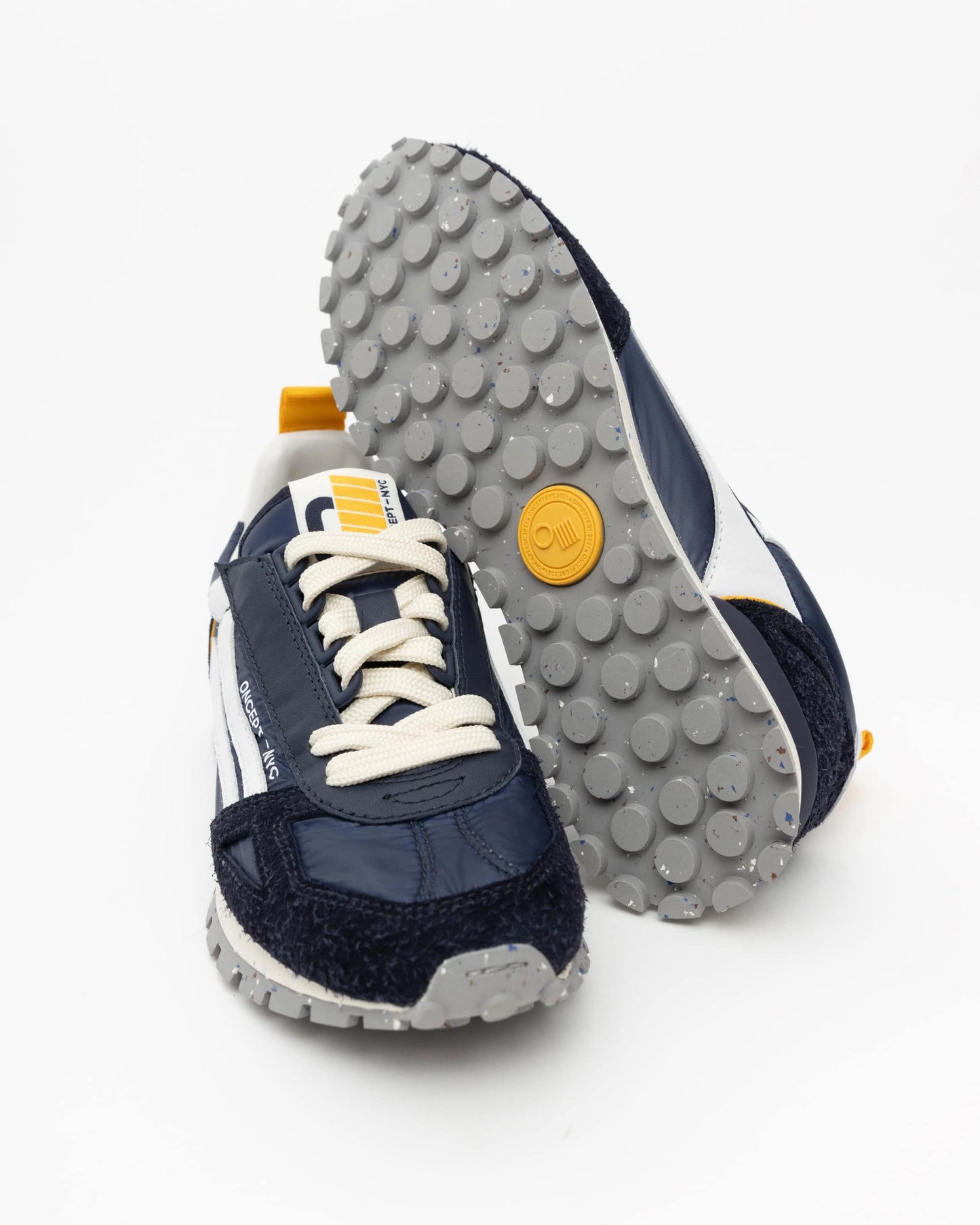 Introducing the Toronto Shoe by Oncept: a stylish pair of navy and white sneakers featuring lightweight, textured gray soles. One shoe stands upright while the other lies on its side, highlighting a wavy sole design and yellow accents on both the tongue and heel.