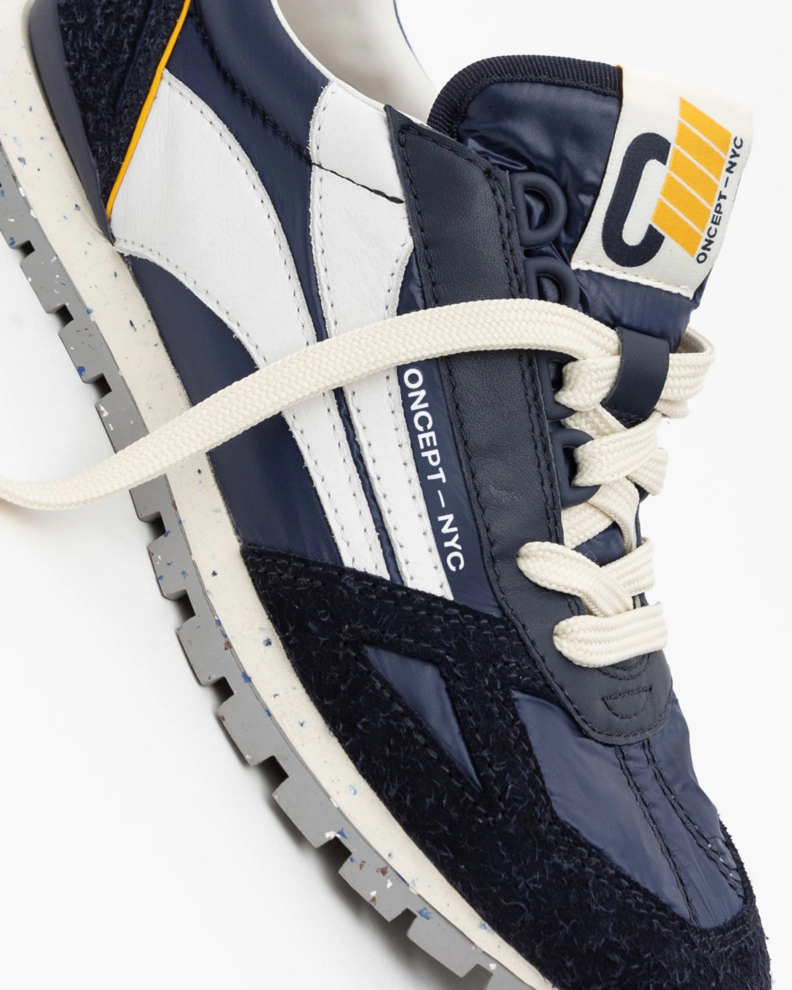 Close-up of the Toronto Shoe by Oncept, a stylish navy and white sneaker with a gray speckled sole. The lightweight shoe features "ONCEPT NYC" branding, yellow accents, and is laced with off-white shoelaces.