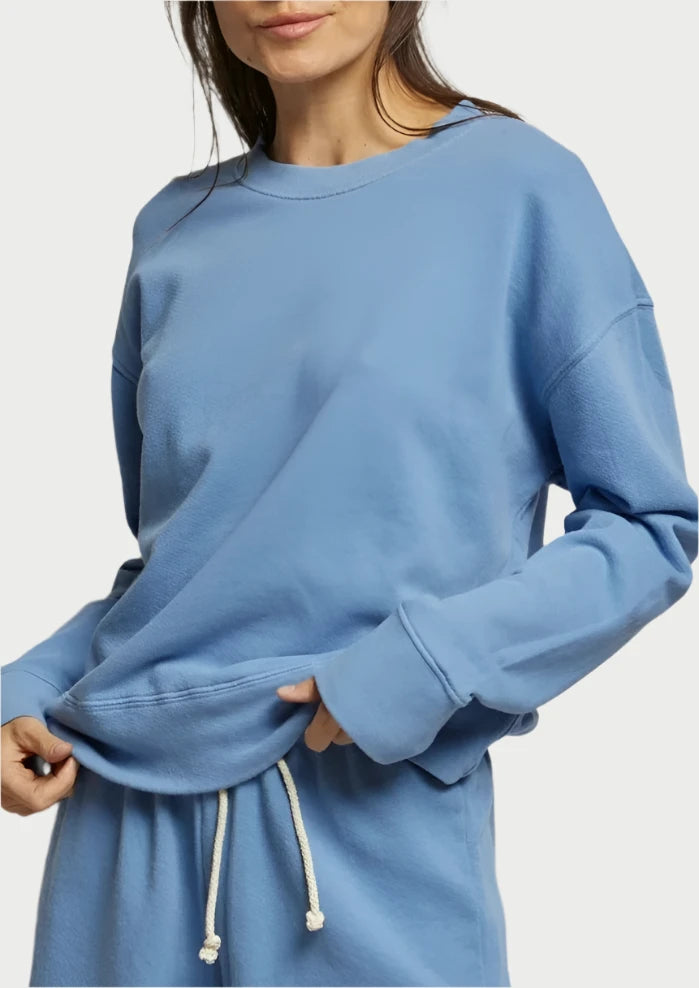 A person with long hair, wearing the Carolina blue Tyler sweatshirt from perfectwhitetee along with matching relaxed fit pants, stands in front of a plain light-colored background. They are slightly smiling and holding the hem of their drop shoulder design sweatshirt with one hand.