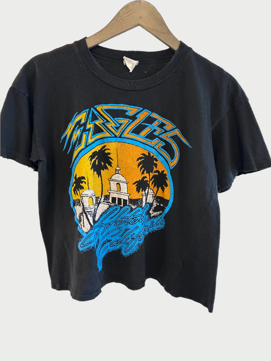 The Eagles Crop Tee by Madeworn features a unique, colorful design of palm trees and buildings against an orange sunset. This black vintage-fit t-shirt showcases blue and orange text that says "Eagles.