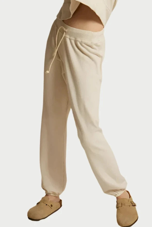 A person wearing the Toni Jogger from perfectwhitetee stands on a beige fabric surface. The cream-colored loungewear set, featuring jogger-style sweatpants with a drawstring waist and a matching top, is paired with beige slip-on shoes with buckles. The individual's head and upper torso are not visible.