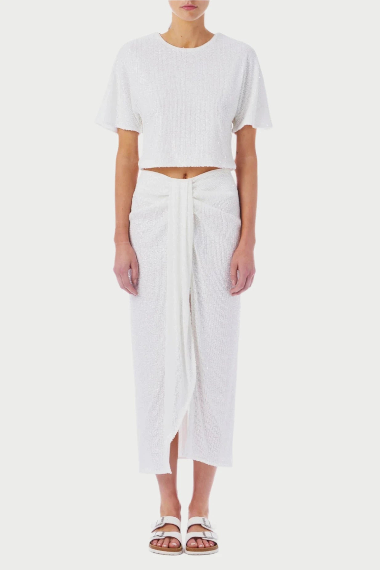 A person stands against a white background, wearing the "Let's Elope Parreo" from Le Superbe—a white short-sleeve crop top paired with a high-waisted midi skirt featuring a front knot detail. The outfit, perfect for a Summer White Party or Wedding Weekend, is complemented by white sandals. Simple and elegant.