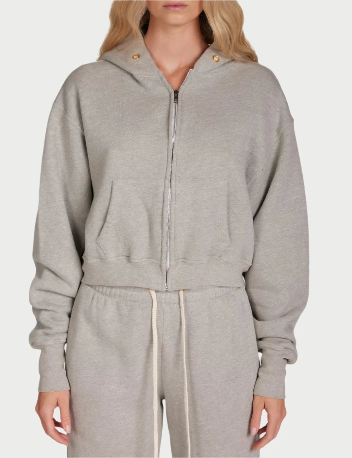 A woman with long, wavy blonde hair is standing against a plain white background. She is wearing the Zendaya Crop Zip Hoodie in light gray by Les Tien, featuring gold-plated grommets and matching sweatpants made from 100% cotton fleece. Her expression is neutral as she faces the camera directly.