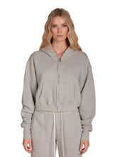 Load image into Gallery viewer, A woman with long, wavy blonde hair is standing against a plain white background. She is wearing the Zendaya Crop Zip Hoodie in light gray by Les Tien, featuring gold-plated grommets and matching sweatpants made from 100% cotton fleece. Her expression is neutral as she faces the camera directly.