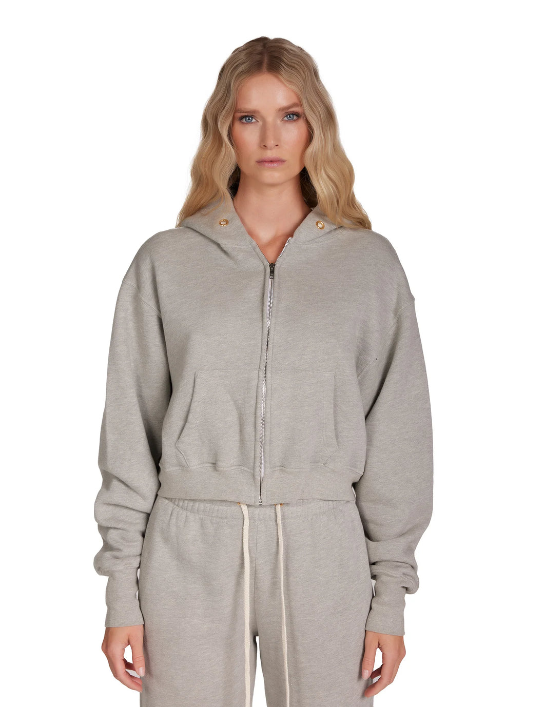 A woman with long, wavy blonde hair is standing against a plain white background. She is wearing the Zendaya Crop Zip Hoodie in light gray by Les Tien, featuring gold-plated grommets and matching sweatpants made from 100% cotton fleece. Her expression is neutral as she faces the camera directly.