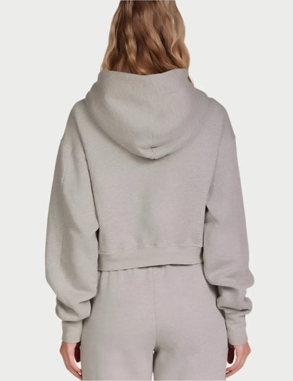 A person with long, wavy hair is standing with their back to the camera, wearing an oversized light gray Zendaya Crop Zip Hoodie by Les Tien and matching high-waisted, wide-leg sweatpants made from 100% cotton fleece. The outfit looks comfortable and casual. The background is plain white.