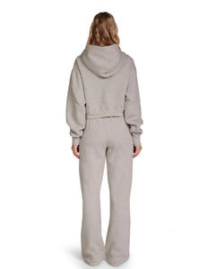 A person with long, wavy hair is standing with their back to the camera, wearing an oversized light gray Zendaya Crop Zip Hoodie by Les Tien and matching high-waisted, wide-leg sweatpants made from 100% cotton fleece. The outfit looks comfortable and casual. The background is plain white.