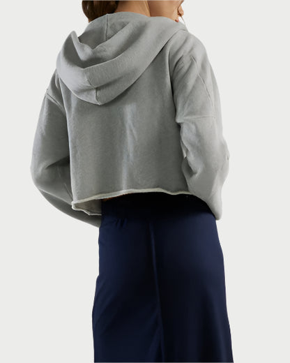 The individual dons a cropped light gray Rosa Raw Zip Hoodie by Les Tien with a long, straight dark blue skirt. The ensemble's elegant simplicity is accentuated by the plain, light-colored background.